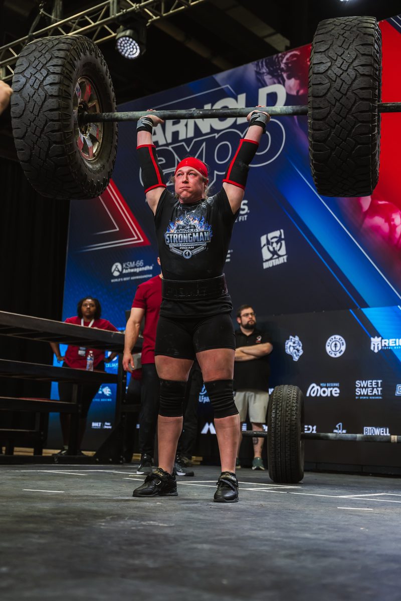 Sarah Johnson - 2026 Arnold Pro/Am U73kg World Champion - pressing a barbell loaded with tires at the Arnold Expo